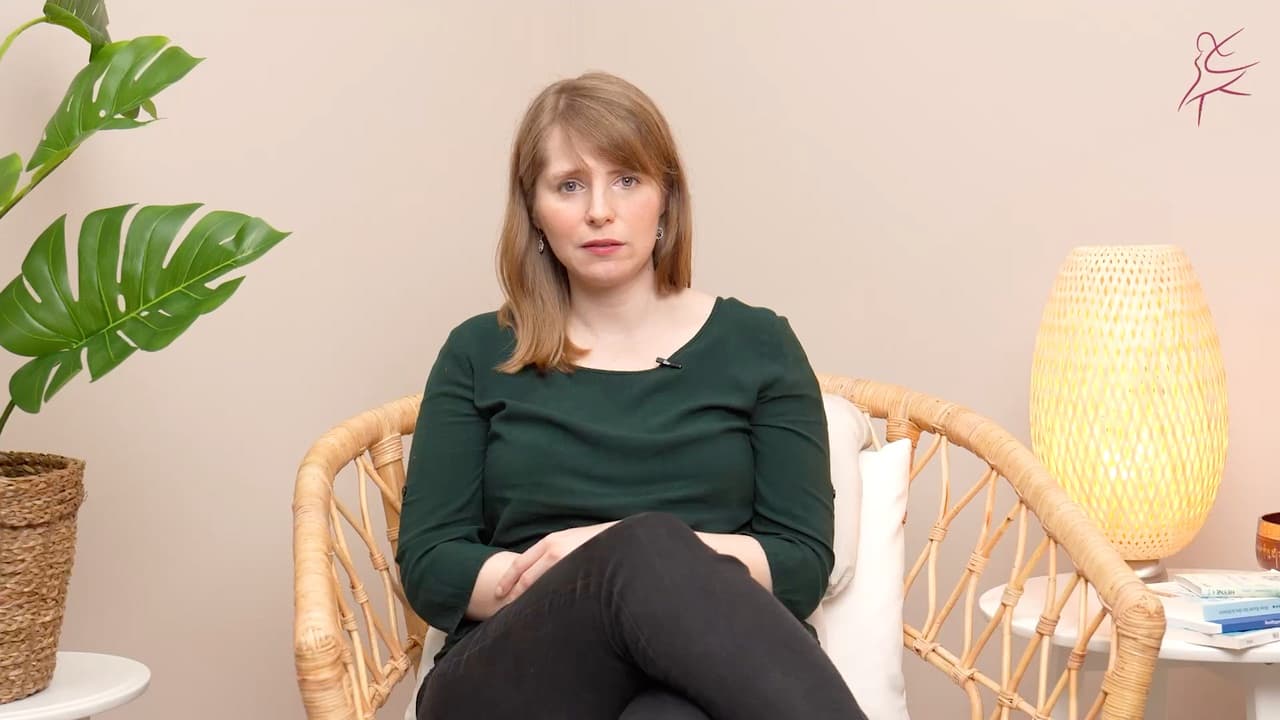 Eine Frau sitzt in einem Korbsessel in einem hellen Raum und spricht in die Kamera