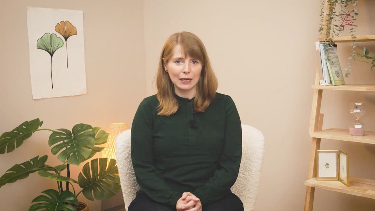 Eine Frau mit rotblonden Haaren sitzt vor einem Bild mit Ginkgoblättern und spricht in die Kamera